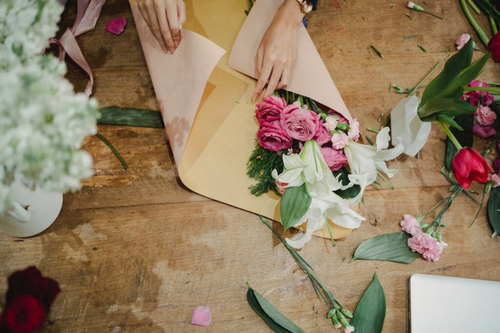 Crop faceless female florist holding craft paper and flowers and creating bouquet in floral shop